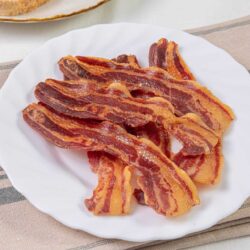 Crispy bacon strips on a white plate with baked Bree background.
