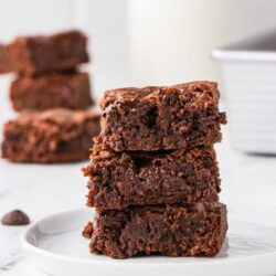 Fudgy triple chocolate brownies stacked on a white plate with chocolate chips scattered around.