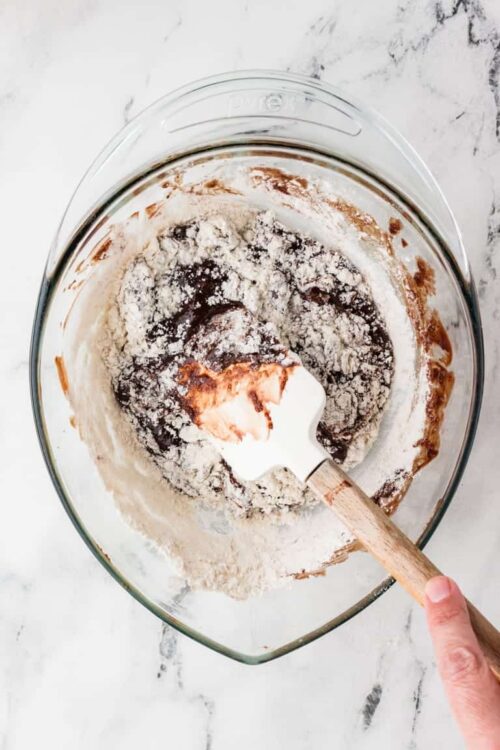 Chocolate cookie dough being mixed in a glass bowl with a spatula on a white marble surface.