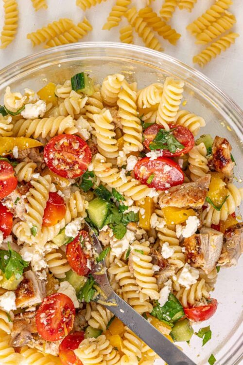 Fusilli pasta salad with cherry tomatoes, cucumbers, cooked chicken, feta cheese, and fresh herbs in a glass bowl.