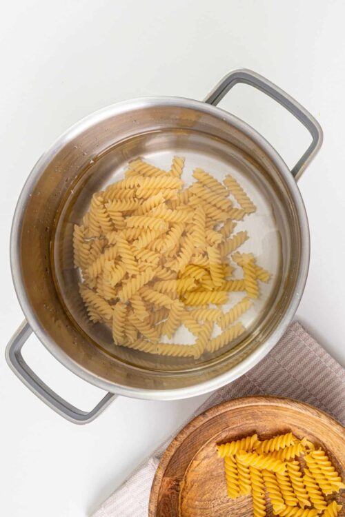 Uncooked fusilli pasta boiling in a stainless steel pot on a white surface.