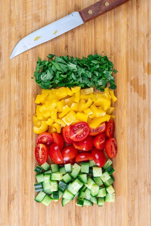 Fresh chopped herbs, yellow bell peppers, cherry tomatoes, and cucumber on a wooden cutting board.