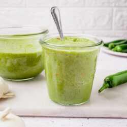 Creamy green jalapeño cilantro dressing in glass jars with fresh garlic and jalapeño peppers on white surface - Baked Bree.