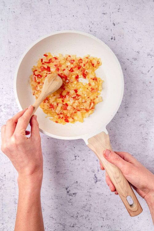 Diced pineapple and red bell peppers cooking in a white skillet with wooden handle.