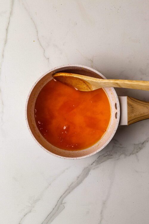 Sweet homemade tomato soup in a speckled pink bowl with a wooden spoon on a white marble surface.