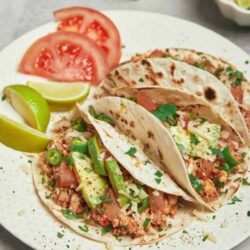 Toasted soft tacos with seasoned ground beef, fresh vegetables, and herbs on a white plate.