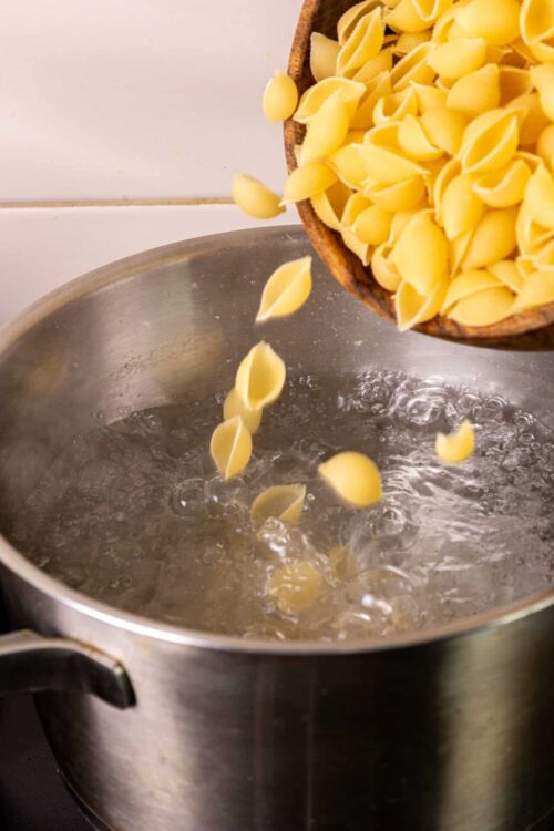 Dried pasta being poured into boiling water in a stainless steel pot.