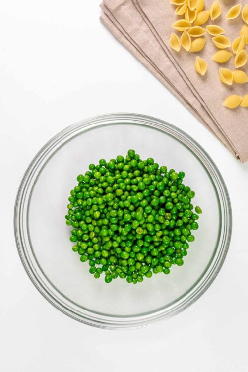 Fresh green peas in a glass bowl on a white surface with dried pasta and a beige napkin.