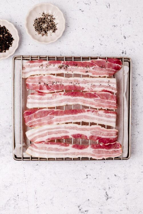 Sliced raw bacon on a wire rack with salt and pepper, ready for cooking or baking.