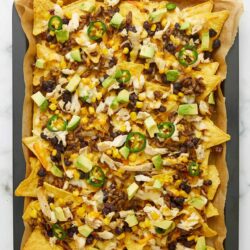 Crispy nachos with chicken, cheese, black beans, corn, jalapenos, and green onions on baking sheet.