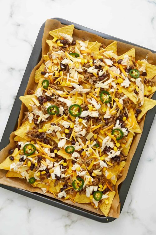 Crispy sheet pan nachos topped with melted cheese, shredded chicken, ground beef, jalapeños, corn, and cilantro.