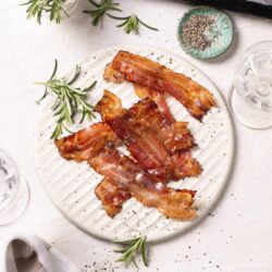 Crispy bacon strips on a white plate with fresh rosemary and black pepper, styled for a savory breakfast or brunch.