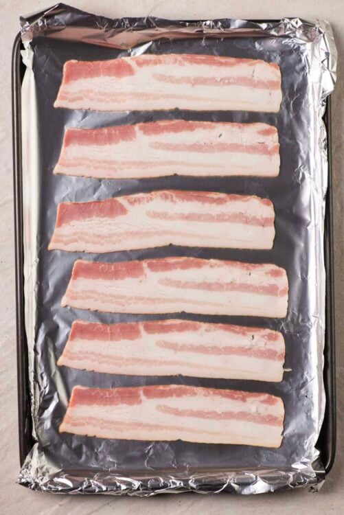 Uncooked bacon slices on foil-lined baking sheet for baking or frying.
