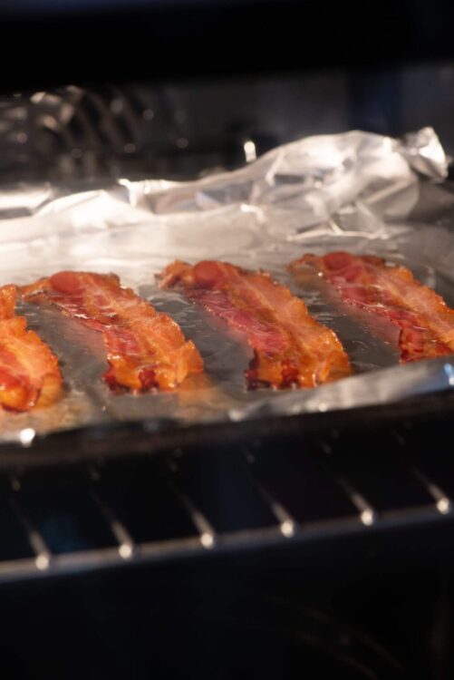 Crispy bacon strips cooking on aluminum foil inside an oven.