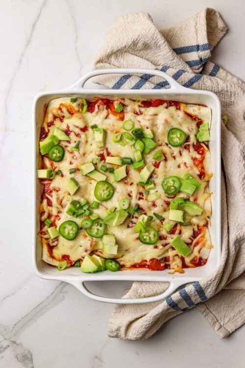 Baked enchiladas with melted cheese, green onions, and avocado slices in a white baking dish.