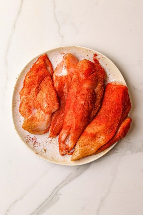 Seasoned raw chicken breasts on a white plate for baking or grilling.