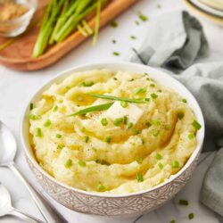 Creamy mashed potatoes with butter and green onions in a white bowl, served with herbs and side dishes.