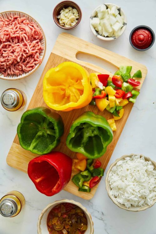 Fresh colorful stuffed bell peppers with chopped vegetables and ground meat ingredients on a wooden cutting board.