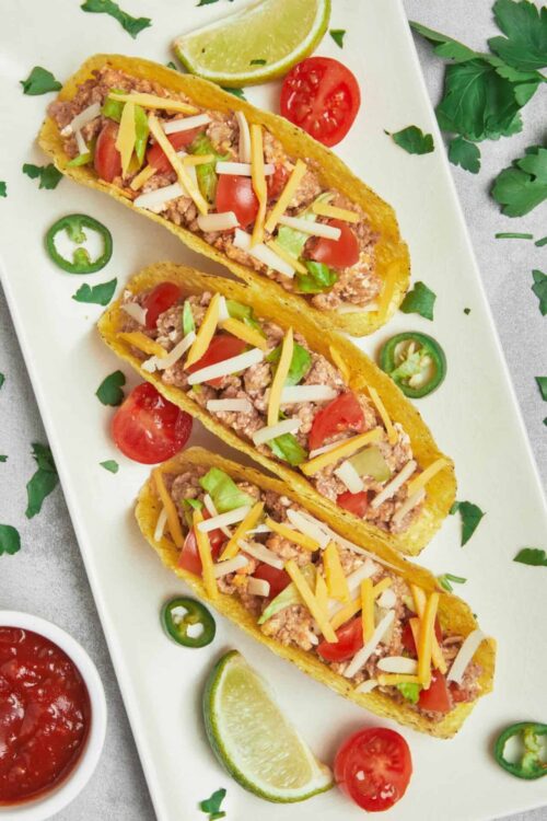 Fresh beef taco boats with shredded cheese, tomatoes, lettuce, lime, and jalapeños on a white platter.