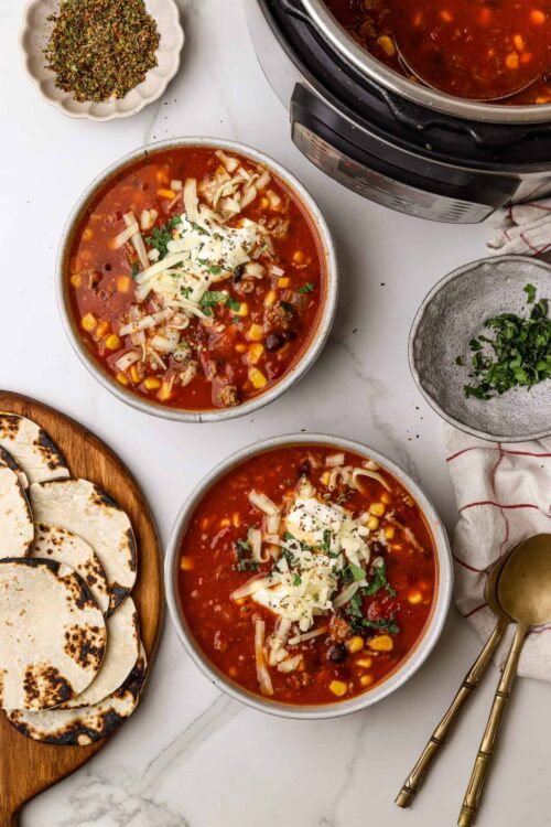 Cheesy vegetable chili in bowls with shredded cheese, fresh herbs, and baked tortillas on a wooden platter.