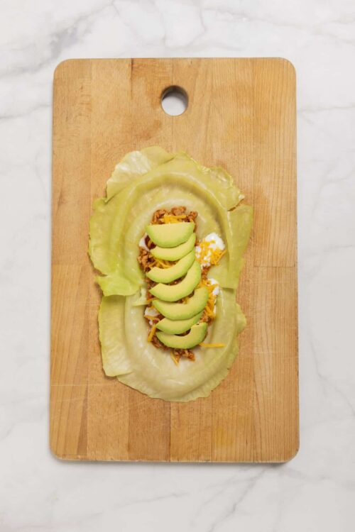 Savory lettuce wrap with ground meat, shredded cheese, avocado slices, and a boiled egg on a wooden cutting board.