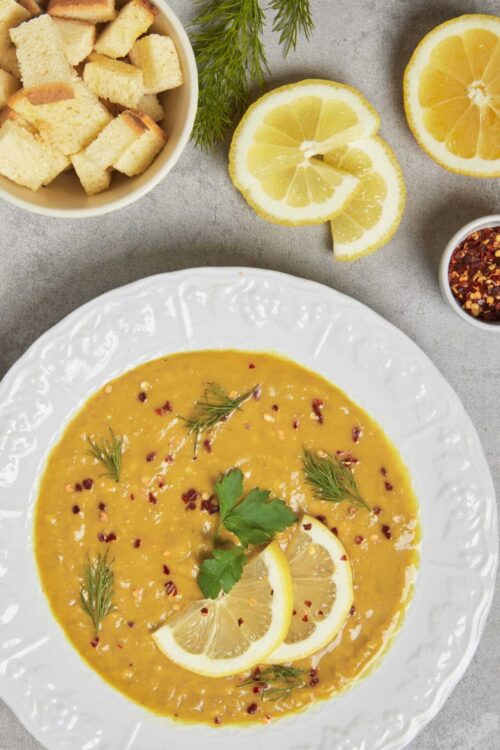 Creamy lemon soup garnished with herbs and red pepper flakes, served on a white plate with lemon slices and bread cubes.
