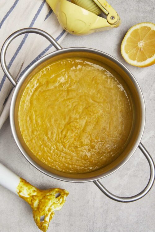 Lemon curd in a stainless steel saucepan with fresh lemon slices and a lemon juicer on a textured surface.