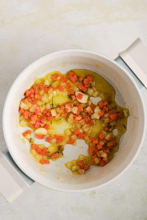 Diced vegetables sautéing in olive oil inside a white skillet.