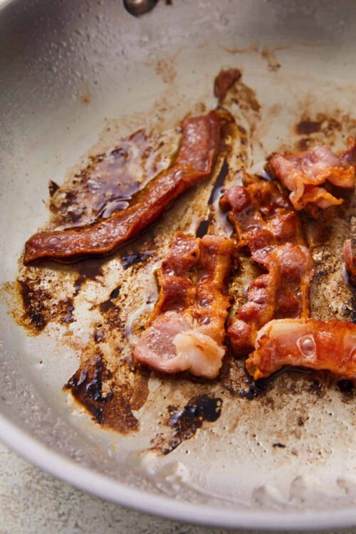 Crispy bacon slices cooking in a skillet with caramelized edges and rendered fat.