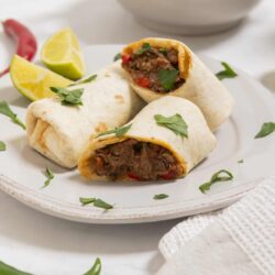 Savory beef burritos on white plate with lime wedges and green chili, Mexican-inspired comfort food, bakedbree.com.