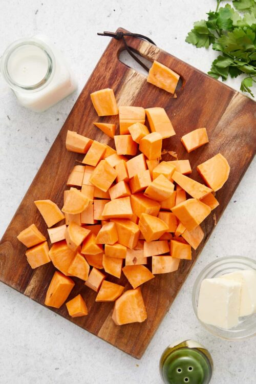 Cubed sweet potatoes on a wooden cutting board with milk, butter, and fresh herbs.