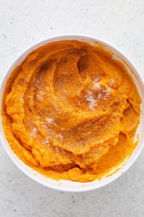 Creamy mashed sweet potatoes in a white bowl topped with black pepper and sea salt.