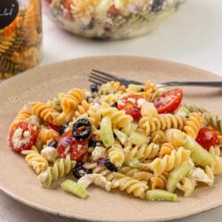 Creamy pasta salad with cherry tomatoes, olives, and feta cheese on a beige plate.