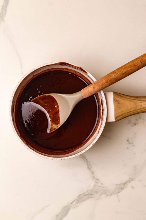 Rich chocolate sauce in a white saucepan with a wooden spoon, on a white marble surface.