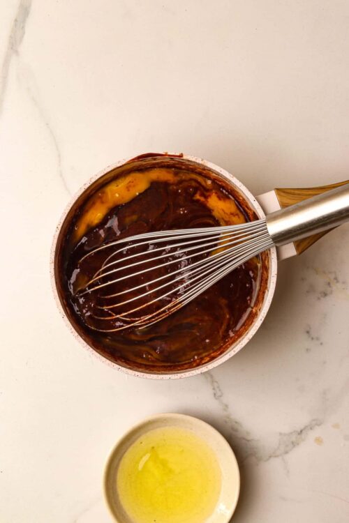 Rich chocolate sauce in a bowl with a stainless steel whisk on a marble countertop.