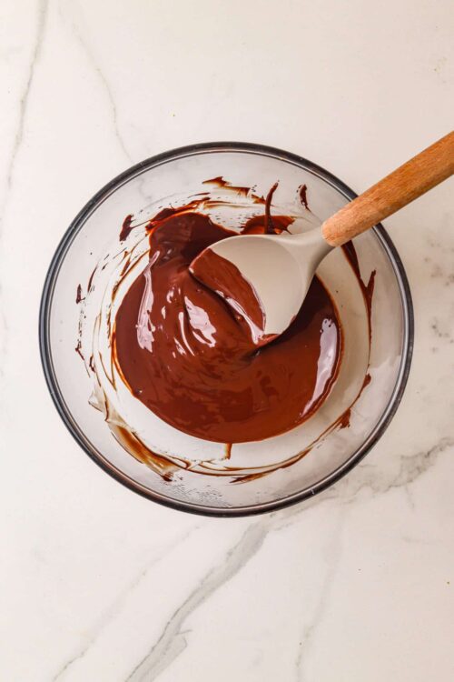 Rich melted chocolate in a glass bowl with a white spatula on a marble countertop.