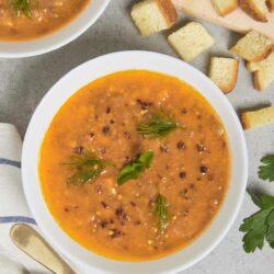 Butternut squash soup with fresh herbs and black pepper, served in a white bowl, with toasted bread slices on a wooden cutting board.