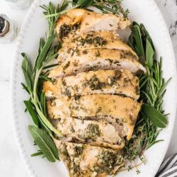 Roasted herb chicken breast on white platter with rosemary and sage sprigs.