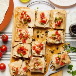 Cheesy vegetable taquitos with salsa on wooden cutting board, fresh tomatoes, and olives.