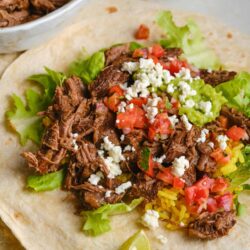 Tender shredded beef barbacoa on a soft corn tortilla with fresh lettuce, tomatoes, crumbled cheese, and lime wedges.