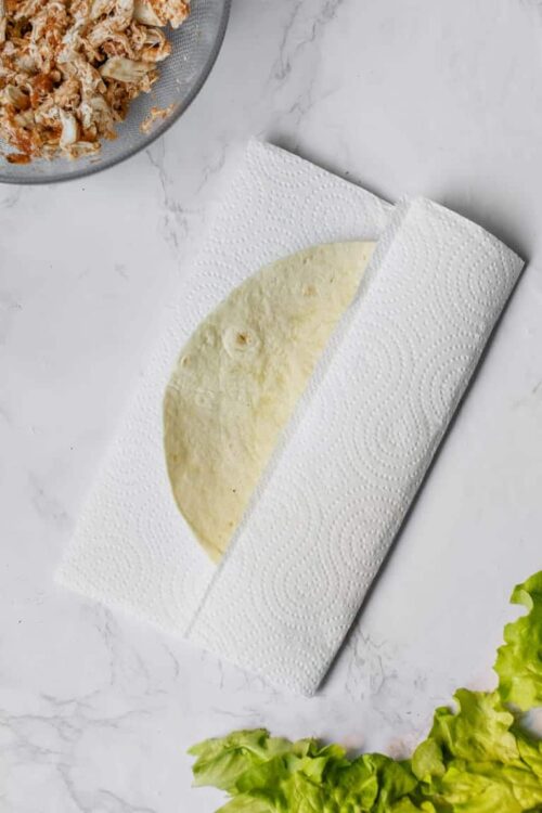 Flour tortilla resting on paper towel with shredded chicken in a bowl nearby.