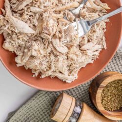 Shredded cooked chicken in a rustic bowl with spices on a textured cloth.