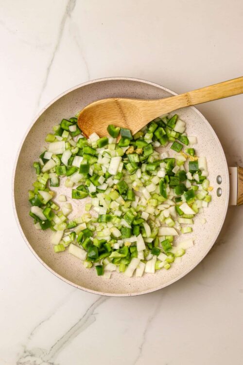 Diced green onions cooking in a skillet with a wooden spoon on a white marble countertop.