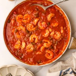 Shrimp and tomato stew in a white skillet with a silver spoon and fresh herbs in the background.