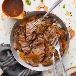 Slow cooker beef roast with gravy and fresh herbs on a white surface.