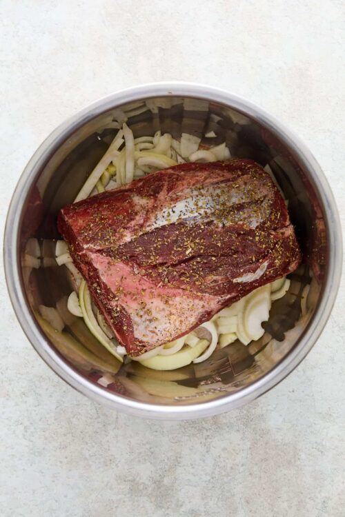 Seasoned beef roast topped with spices over sliced onions in a slow cooker.
