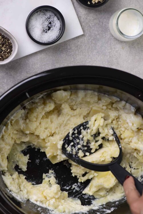 Buttery mashed potatoes being prepared in a slow cooker with a black plastic masher.