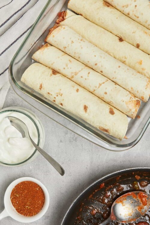 Savory rolled tortillas in a glass baking dish with chili sauce and creamy sour cream on the side.
