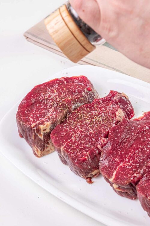 Sliced raw beef steaks seasoned with salt and pepper on a white platter.