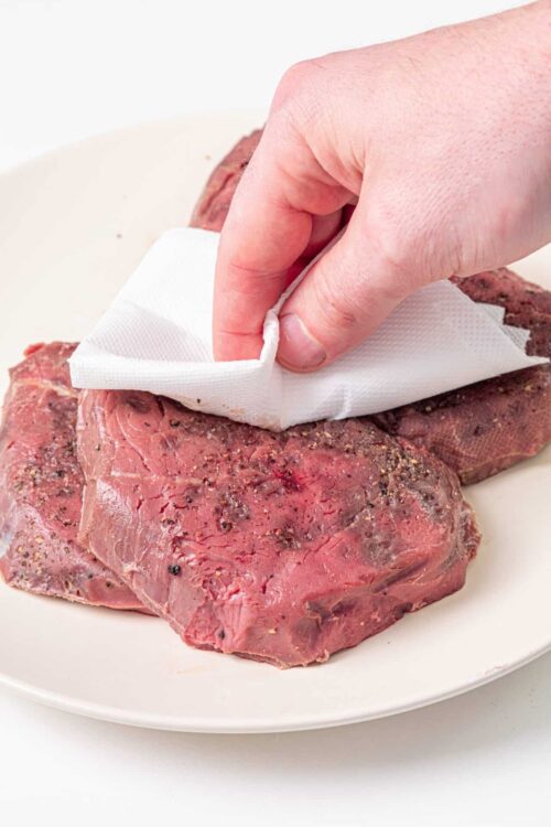 Juicy raw steak being seasoned and prepared for cooking on a white plate.